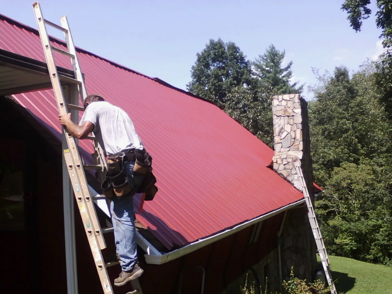 Roofer on a ladder beside a red metal roof and stone chimney — Soffit & Fascia Repair by Roofing Brothers in Jackson Heights